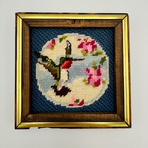 May include: A framed needlepoint embroidery depicting a hummingbird in flight with pink flowers on a blue background.
