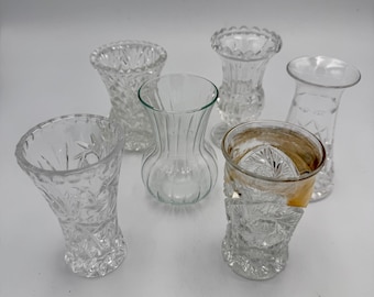 May include: A collection of six clear glass vases of varying shapes and sizes. Four vases feature intricate cut-glass patterns, one has vertical ribbing, and one is plain with a smooth rim. The items are arranged in a group on a neutral surface.