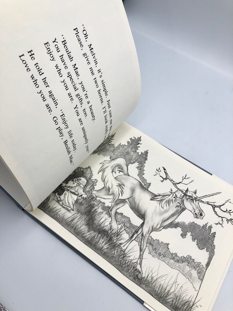 May include: A black and white illustration of a unicorn with a branch for a horn, running through a field of grass. A small figure in a dress is running behind the unicorn. The text reads: "Oh, Melvin, it's simple. Just cast me those two horns. I'll look really beautiful. Beulah Mae, you're a beauty. You have special gifts, too. Enjoy who you are. You are uniquely you. He told her again, "Enjoy life today. Love who you are. Go play, Beulah Mae."