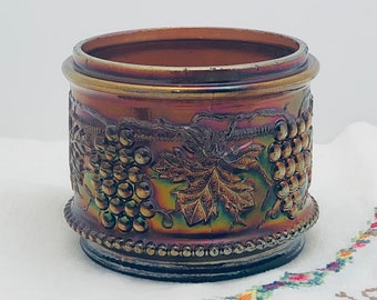 May include: A vintage iridescent glass jar with a grapevine design. The jar is a deep purple color with a brown rim and a beaded edge.