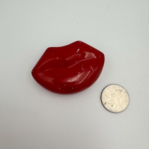 Pode incluir: Um objeto brilhante em forma de lábios vermelhos ao lado de uma moeda de um quarto de dólar dos EUA. A escultura de lábios é de um vermelho vibrante com uma superfície lisa e reflexiva. A moeda é prateada e mostra o perfil de George Washington.