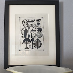 May include: A framed black and white cross-stitch artwork with kitchen-themed illustrations, including a watermelon slice, fish, and pineapple. Below, a yellow Pyrex bowl with black kitchen-themed designs.
