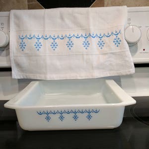 May include: White rectangular baking dish and matching kitchen towel with blue snowflake and scalloped border design. The towel is hanging on a white stove. The baking dish is sitting on a black stove top.