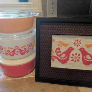 May include: A stack of vintage Pyrex bowls in orange, white, and red, each with a clear glass lid. The white bowl features a pink and orange bird and floral design. A framed cross-stitch with a bird and floral pattern is also visible.