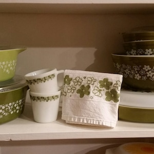 Op de afbeelding: Een verzameling vintage keukengerei, waaronder groene Pyrex-kommen en -schalen met witte bloempatronen. Twee witte mokken met groene bloemaccenten en een witte handdoek met groene bloemborduurwerk zijn ook aanwezig.