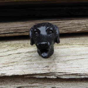 May include: Black plastic dog head figurine. The dog has a black nose, black eyes, and a black tongue sticking out. The figurine is on a wooden surface.