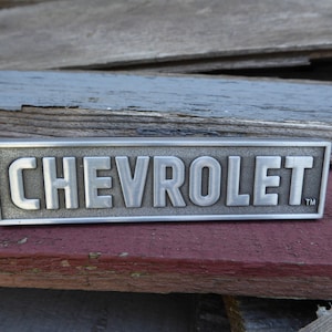 Könnte beinhalten: Ein silbernes Metallemblem mit dem Wort "CHEVROLET" in erhabenen Buchstaben und einem Warenzeichen.