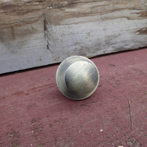 Op de afbeelding: Een ronde, messingkleurige kastknop met een licht verhoogd, getextureerd oppervlak. De knop heeft een verweerde, antieke afwerking.