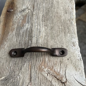 Può includere: Una maniglia per cassetto in metallo marrone scuro con forma arrotondata. La maniglia è montata su una superficie in legno invecchiato.