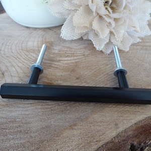 Peut inclure: Une poignée de meuble noire avec une barre rectangulaire et deux vis argentées. La quincaillerie est posée sur un fond en bois naturel, avec un vase blanc et une fleur beige en arrière-plan.