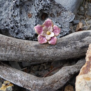 May include: A pink and white enamel flower brooch with gold accents. The brooch is sitting on a piece of driftwood.