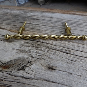 May include: A gold-toned twisted metal drawer pull with rounded ends. The pull is designed for use on cabinets, drawers, or furniture.