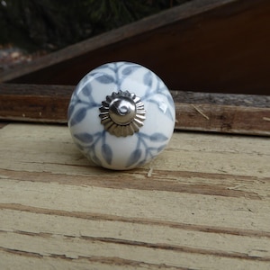 May include: A round, white ceramic cabinet knob with a silver-colored metal base. The knob features a blue leaf pattern and is set against a rustic wooden background. This decorative hardware is suitable for drawers and cabinets.