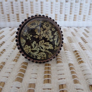 Könnte beinhalten: Ein Ring mit schwarzem und goldenem Blumenmuster und einem runden Glascabochon.