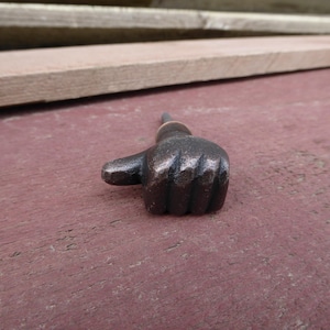 May include: A black metal thumbs up door knob or pull. The knob is in the shape of a hand with the thumb pointing up.