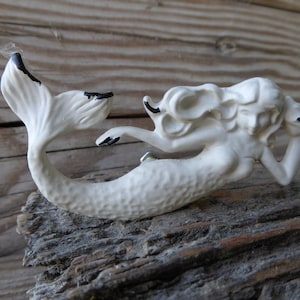 Könnte beinhalten: Weiße Keramik-Meerjungfrauenfigur mit schwarzen Akzenten. Die Meerjungfrau liegt auf der Seite, ihr Schwanz ist hinter ihr eingerollt.