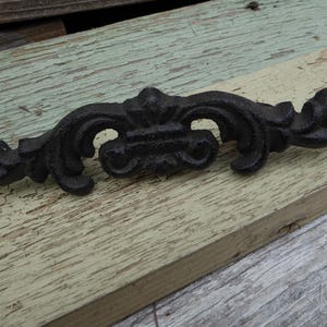 May include: A black, decorative cast iron drawer pull with an elaborate, symmetrical design. The pull features scrolled and floral details, and is mounted on a weathered, light-coloured wooden surface. Suitable for furniture restoration or decorative purposes.
