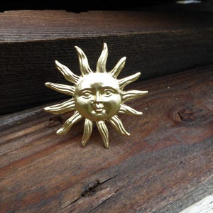 May include: A gold-colored sun-shaped brooch with a smiling face and radiating rays. The brooch is made of metal and is set against a weathered wooden surface. The sun design is a classic and timeless symbol.