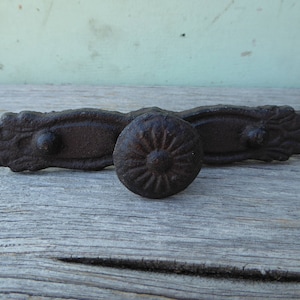May include: A set of antique-style, cast iron hardware. The set includes a decorative pull with floral accents and a round knob with a sunburst design. The hardware is finished in a dark, rustic brown.