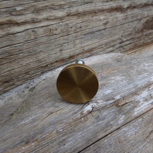 Op de afbeelding: Een messingkleurige, ronde kastknop met een geborstelde afwerking. De knop heeft een kleine, zilverkleurige steel. De knop wordt getoond tegen een verweerde, grijze houten achtergrond. Deze hardware is geschikt voor laden en kasten.
