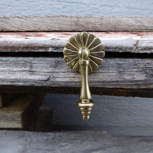 May include: A brass drawer pull with a floral design. The pull features a round, sunburst-like face and a decorative, elongated handle. The hardware is mounted on weathered wooden planks, showcasing its vintage aesthetic.