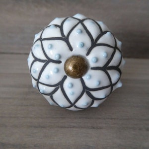 May include: White ceramic cabinet knob with a floral design. The knob features black outlines, small light blue dots, and a gold-colored center. The knob is round and has a textured surface, suitable for furniture or decor.