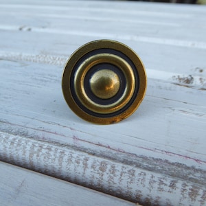 May include: A circular cabinet knob in antique brass, featuring a series of concentric circles. The central button is raised, and the finish has a warm, aged brass tone. This knob would suit vintage or rustic furniture, with a diameter of approximately 3.8 cm.