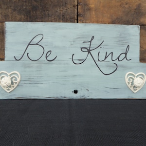 Puede incluir: Un letrero de madera azul claro con las palabras "Be Kind" escritas en letra negra. El letrero tiene dos corazones decorativos blancos a los lados.