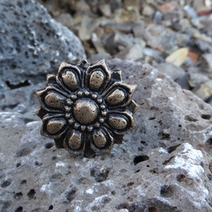 May include: A decorative metal flower with a gold finish. The flower has eight petals and a center with a raised circle. The flower is sitting on a gray rock.