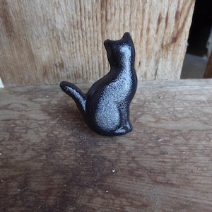 May include: A small, black cat figurine with a textured surface. The cat is sitting upright with its tail curved to the side. The figurine is placed on a wooden surface with a natural wood grain.