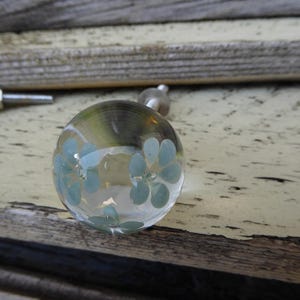May include: A spherical clear glass cabinet knob featuring an embedded light blue floral pattern with five petals. The knob is attached to a silver-toned metal bolt extending from the back, designed for installation on furniture or doors.