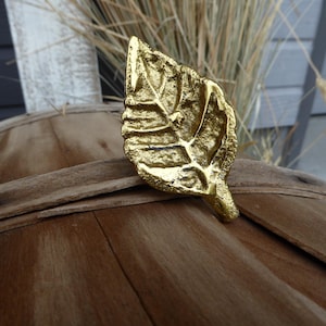 May include: A gold-colored leaf-shaped decorative object. The leaf has detailed veining and a textured surface. It is placed on a wooden surface, with dried grass in the background. The object appears to be a decorative accent.