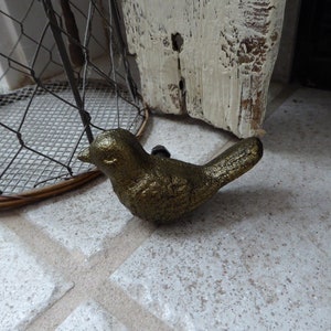 May include: A gold-colored bird-shaped cabinet knob. The decorative hardware piece is made of metal and has a textured surface. The bird is positioned on a light-colored tiled floor. A wire mesh object is in the background.