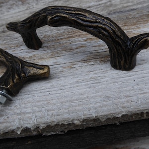 May include: Two bronze-colored cabinet pulls in the shape of tree branches. The pulls have a textured surface and are mounted with a bolt, washer, and nut. The hardware is designed to look like a natural branch.