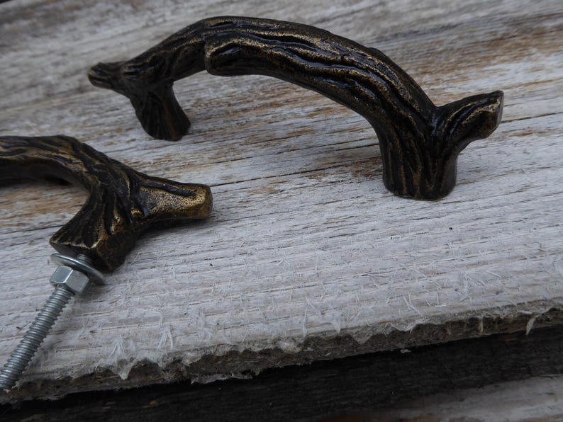 May include: Two bronze-colored cabinet pulls in the shape of tree branches. The pulls have a textured surface and are mounted with a bolt, washer, and nut. The hardware is designed to look like a natural branch.