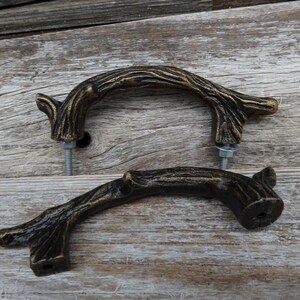 May include: Two bronze-colored, branch-shaped cabinet pulls with mounting hardware. The pulls have a textured, naturalistic design, suggesting a rustic or woodland theme. They are set against a weathered wood background.