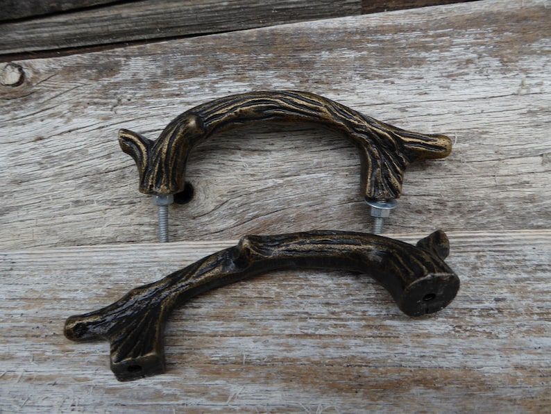 May include: Two bronze-colored, branch-shaped cabinet pulls with mounting hardware. The pulls have a textured, naturalistic design, suggesting a rustic or woodland theme. They are set against a weathered wood background.
