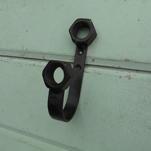 May include: Black metal hook with two hexagonal nuts at the top. The hook is mounted on a light green wall.