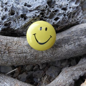 May include: A yellow smiley face button with a black outline. The button is sitting on a piece of weathered wood.
