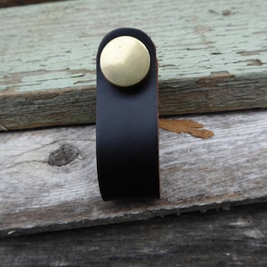 May include: A black leather cabinet pull with a brass-colored circular rivet. The pull is a simple loop design, suitable for drawers or cabinets. The background is weathered wood.