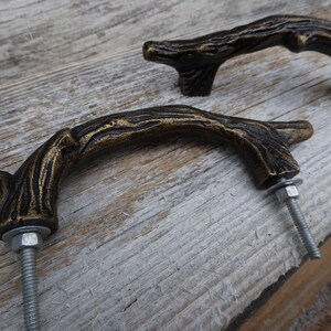 May include: Two bronze-colored, branch-shaped cabinet pulls with a rustic, textured finish. The pulls are mounted with silver bolts and washers, set against a weathered wooden surface. These decorative pulls add a natural, organic touch to furniture.