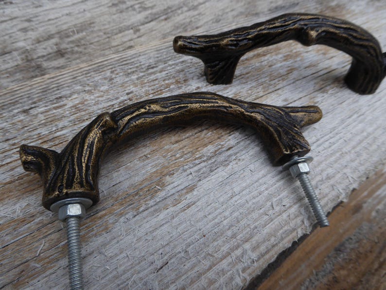May include: Two bronze-colored, branch-shaped cabinet pulls with a rustic, textured finish. The pulls are mounted with silver bolts and washers, set against a weathered wooden surface. These decorative pulls add a natural, organic touch to furniture.