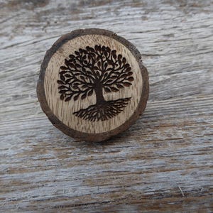 Könnte beinhalten: Ein runder Holz-Anstecker mit einem Lebensbaum-Design. Der Baum ist in das hellfarbene Holz geätzt, mit dunkelbrauner Rinde und Wurzeln. Der Anstecker befindet sich vor einem verwitterten Holz-Hintergrund.