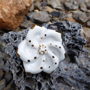 Puede incluir: Pomo de cerámica blanca en forma de flor con detalles dorados y un diseño de espiral. El pomo está sobre una superficie gris texturizada.