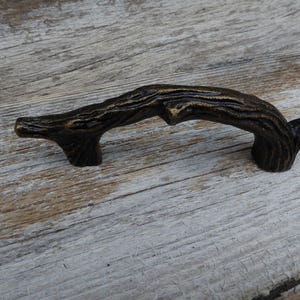 Puede incluir: Tirador de armario de color bronce en forma de rama de árbol. El tirador tiene una superficie texturizada, con detalles que imitan el aspecto natural de la madera. El herraje está diseñado para armarios y cajones.