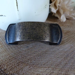 May include: A bronze-coloured cabinet pull with a textured finish. The hardware has a curved, rectangular design with rounded ends. The pull is set against a natural wood background, suggesting a rustic or vintage aesthetic.