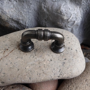 May include: A black metal pipe-style drawer pull with a rounded shape. The pull is sitting on a light gray rock.