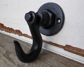 May include: Black metal hook with a round base mounted on a white wall. The hook is in the shape of a large 'J' and is attached to a round metal plate.