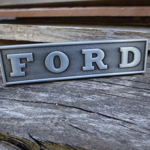 以下が含まれることがあります： 「FORD」という文字が浮き彫りになった銀色の金属製のエンブレムです。