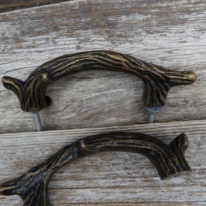 May include: Two bronze-colored, twig-shaped cabinet pulls. The pulls have a textured, bark-like appearance and are mounted with silver screws. The hardware is designed to resemble natural tree branches, adding a rustic touch to furniture.